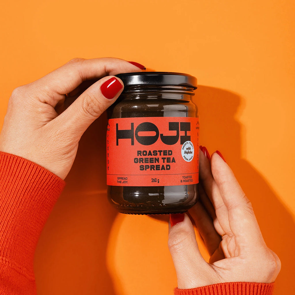 Jar of HOJI Roasted Green Tea Spread held by a person against an orange background