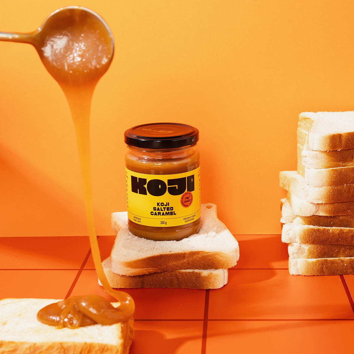 Jar of Koji caramel sauce with a spoonful being poured over toast on an orange background