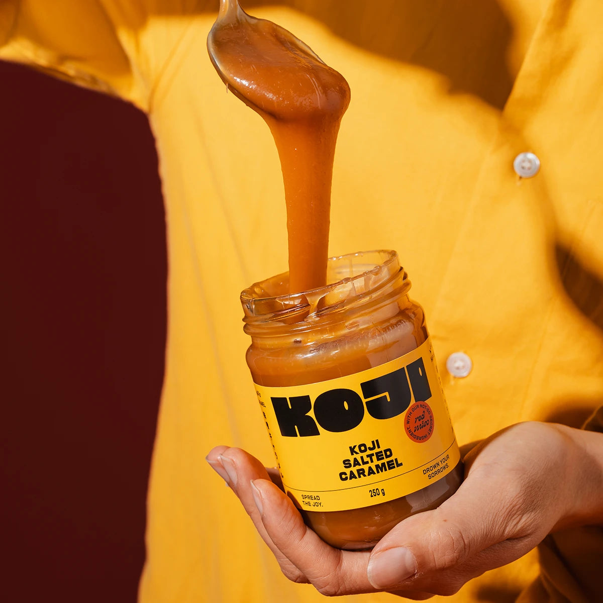 Jar of Koji salted caramel being held with a spoonful of caramel dripping out, person wearing a yellow shirt.