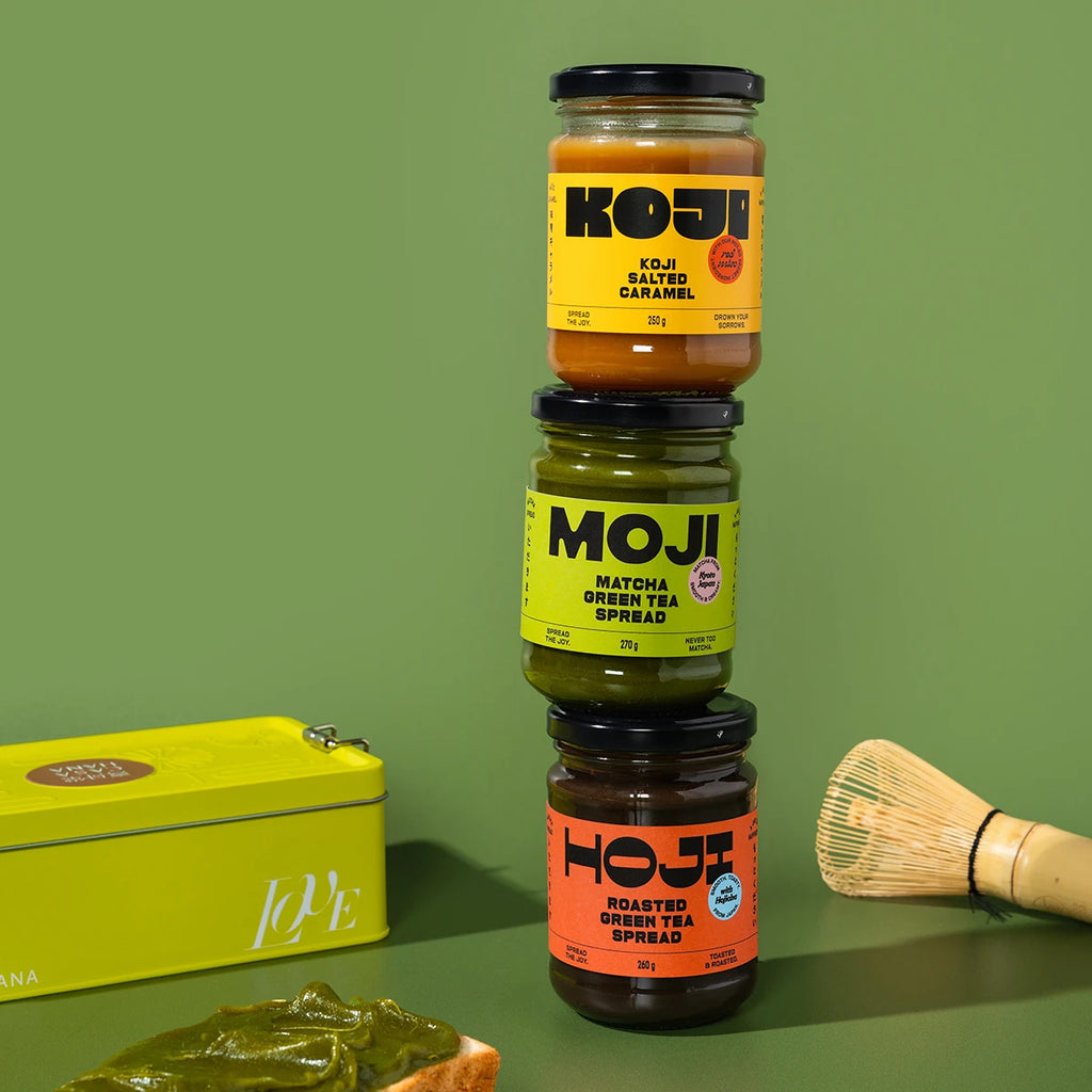 A jar of Koji Salted Caramel, a jar of Moji matcha spread and a jar of Hoji roasted green tea spread stacked on a green background with a small container and wooden tool.