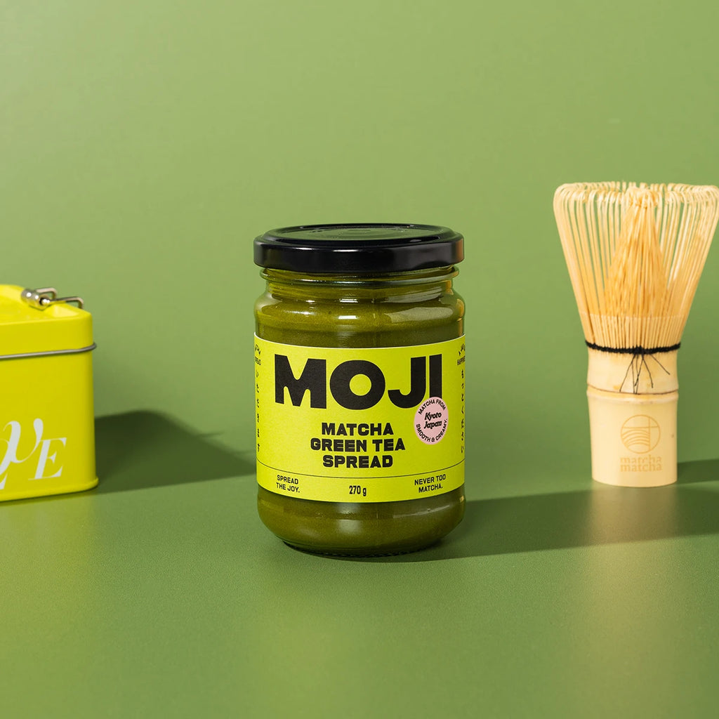 Jar of MOJI Matcha Green Tea Spread with a whisk and container on a green background
