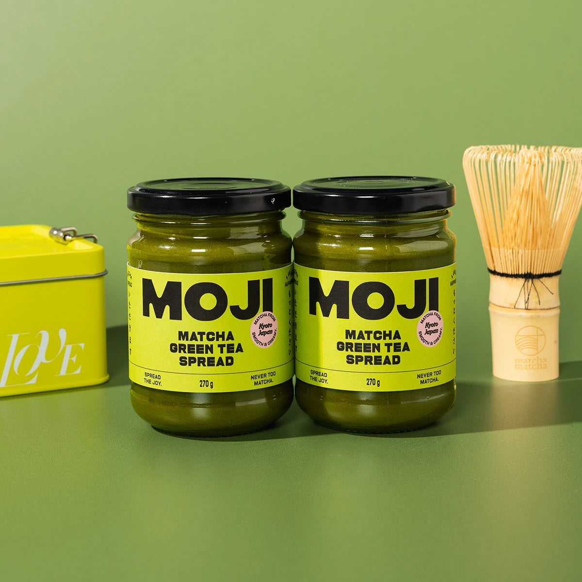 Two jars of MOJI Matcha Green Tea Spread on a green background with a whisk and container.
