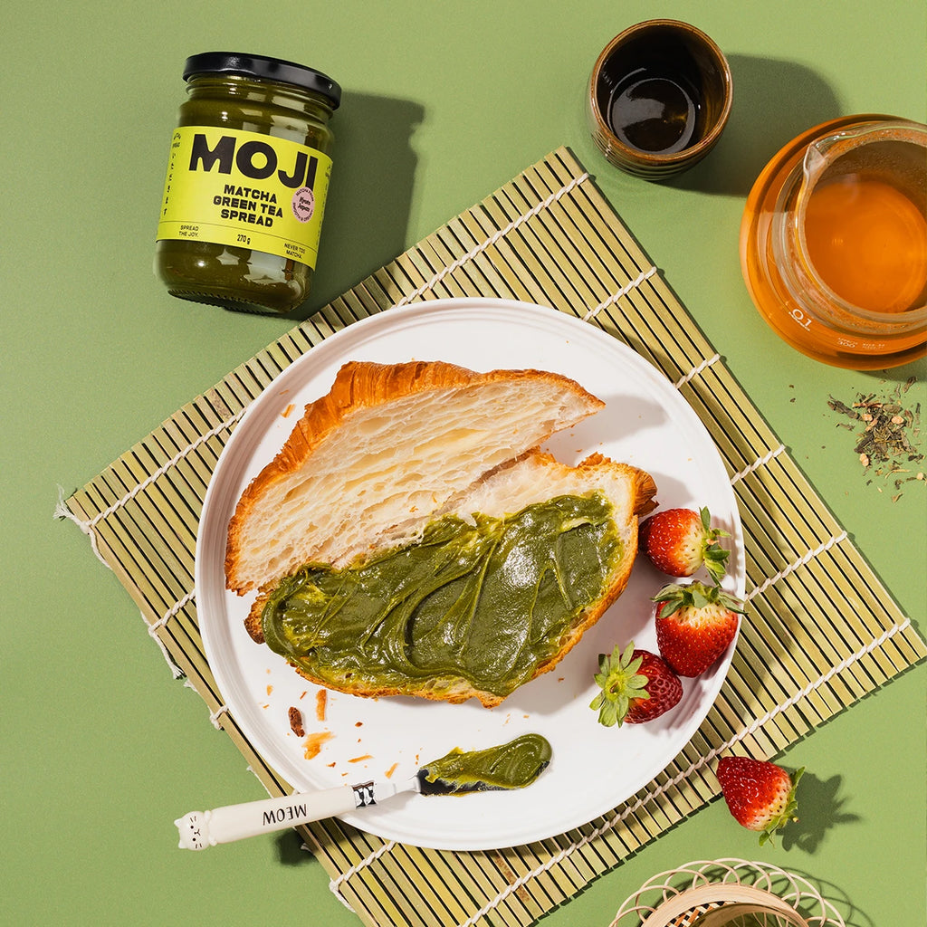 Matcha spread on toast with strawberries, a jar of MOJI Matcha Tea Spread, and a cup of tea on a green background.