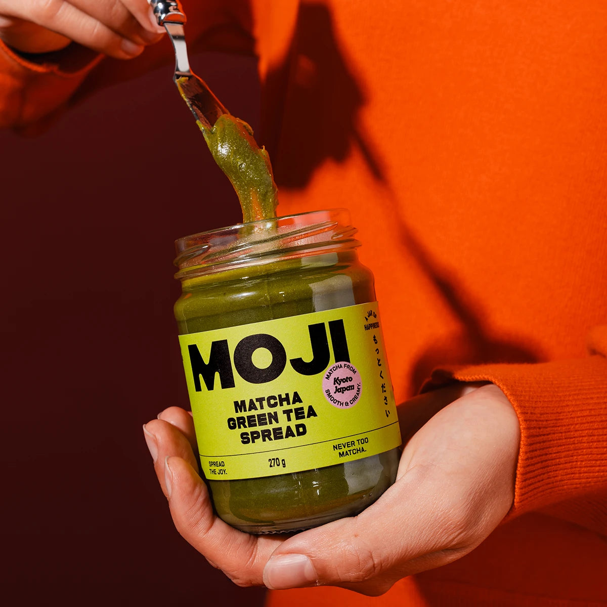 Jar of MOJI Matcha Green Tea Spread with a person spreading it on a spoon against an orange background