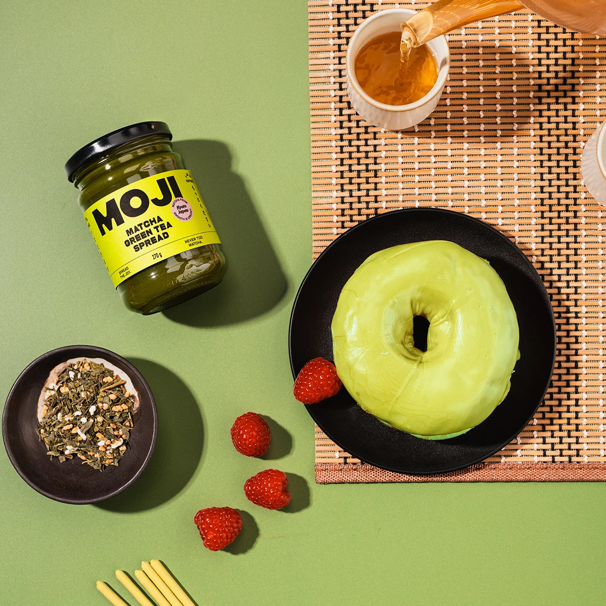 Jar of MOJI Matcha Green Tea Spread on a green background with a green donut and raspberries.